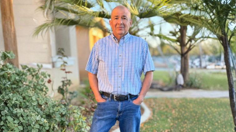 Man in blue button up and jeans stands in front of his house with his hands in his pocket