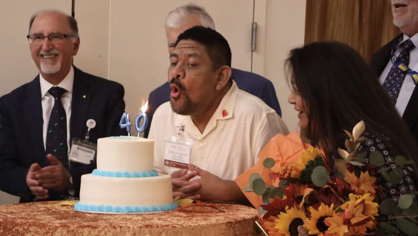 A man blows out candles on a two tiered cake with people surrounding him.
