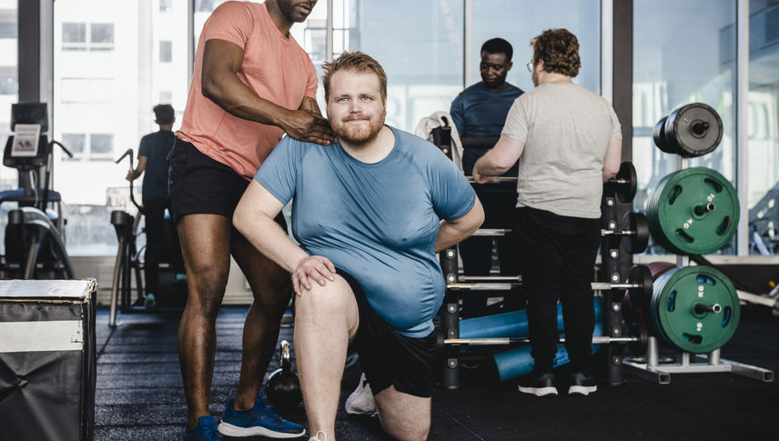 Male fitness instructor assisting overweight man in exercise at gym - stock photo