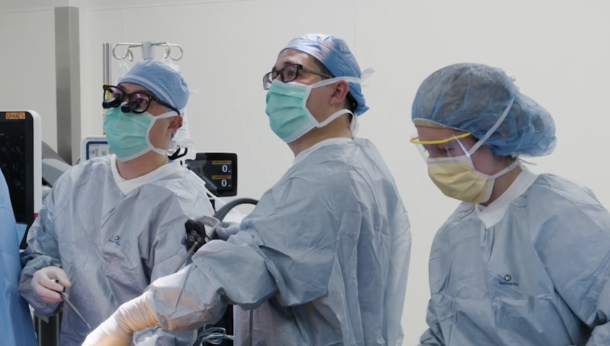 Three people in blue scrubs stand over operating table looking to the viewers left.