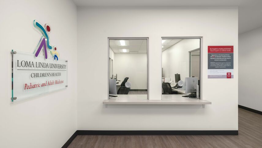Reception area at Loma Linda University Children’s Health Pediatric and Adult Medicine clinic with two service windows, a counter, office workstations visible behind glass, and clinic signage on a bright, modern interior wall.