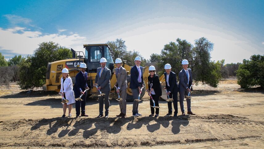 eight hospital leadership representatives dig dirt during the groundbreaking ceremony in front of a dump truck with trees in the background.