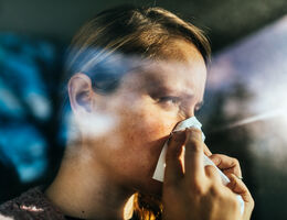 A person holding a tissue to their nose while sitting in a car, with light streaming through the window.