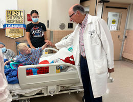young blonde boy sits on hospital gurney in blue hospital gown giving his doctor wearing white coat and glasses a high five with two female medical staff nearby