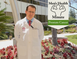 A doctor wearing a white coat stands with his hands behind his back while looking into the camera. He is in front of a medical building