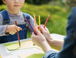 Child holding color pencils and drawing acorss from adult