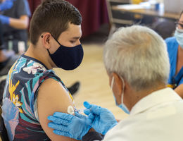 Doctor in white coat gives vaccine to person with short hair and colorful shirt