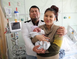 new mother stands with father holding their baby and smiling