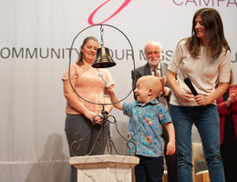 a young Caucasian boy with no hair in s blue shirt and jeans rings a standing bell, smiling with his mom beside him