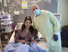 Norah sits in a hospital bed on the left of her father who is wearing a gown and a mask, both smiling at the camera.
