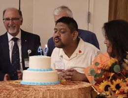 A man blows out candles on a two tiered cake with people surrounding him.