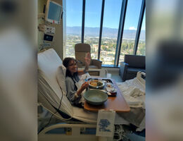 Woman sitting in hospital bed smiling and waving with a large window and scenic view in the background.