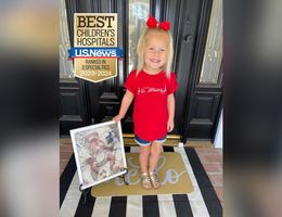 young blonde girl, smiling, holding a framed picture of herself as a baby undergoing treatment