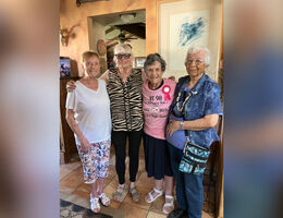 Four elderly women standing shoulder to shoulder smiling. One wears a birthday ribbon pin.