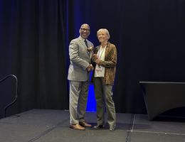 Craig Jackson takes the helm as President of the Association of Schools Advancing Health Professions, receiving the gavel from outgoing President Dr. Deb Larsen Black man in a grey business suit being presented an honorary gavel on stage by a Caucasion women in a pant suit,