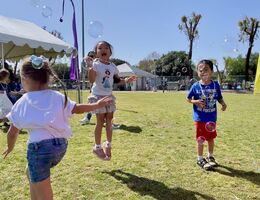 Three children in tee-shirts and shorts play outside in the grass blowing bubbles and smiling 