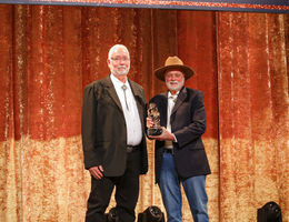 tall Caucasian man with white hair in a suit accepting award on stage from another Caucasian man in a suit and cowboy hat.