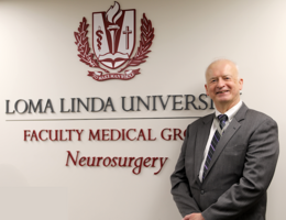 Man stands in front of a white wall with a sign saying Loma Linda University Faculty Medical Group Neurosurgery