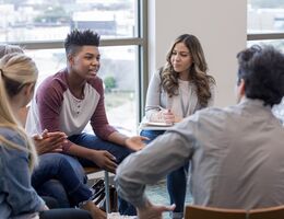 A teen in a red and white shirt sits in a group of other young people during a therapy session during the daytime