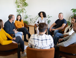 A group of mixed ethnic people sitting in a circle in comfortable chairs holding a support meeting, as they smile and chat with each other