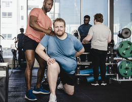 Male fitness instructor assisting overweight man in exercise at gym - stock photo