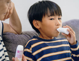 Boy using inhaler while sitting on gray couch, mom next to him supervising