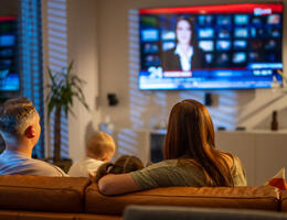 A woman, man, and baby sit on a brown couch and watch the news