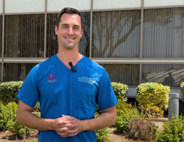 Doctor in blue scrubs stands outside a building surrounded by green bushes