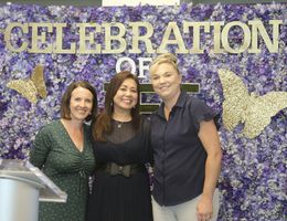 Three women stand smiling in front of purple flower backdrop with gold butterflies4. Sign reads "Celebration of Life"