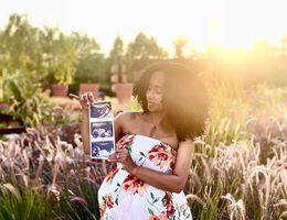 African American woman stands with sonogram in front of field, pregnant and smiling