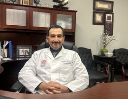 Man sits at desk smiling in white doctors coat.