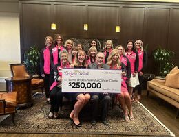 Group of women in pink jackets surrounding woman and man sitting holding oversized check reading a $250,000 donation to the Believe Walk