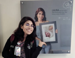 Nurse Laura stands in front of hallway image of herself holding a photo of newborn Laura when she was in NICU