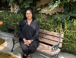 A woman in all black sits on a bench outside in front of beautiful flowers and greenary