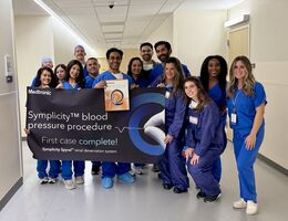 Hospital staff all in blue scrubs stand smiling holding a banner that says "Symplicity blood pressure procedure. First case complete!"