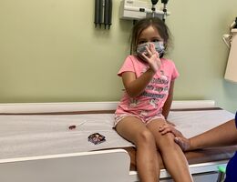 Three-year-old girl sits on exam table in clinic waving to camera about to get vaccination