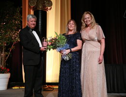 senior vice president for advancement, Rachelle, stands with Don and Penny holding award and flowers smiling for photo on stage