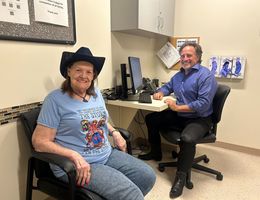 White woman sits smiling at camera in cowgirl cat next to middle aged white male physician. 