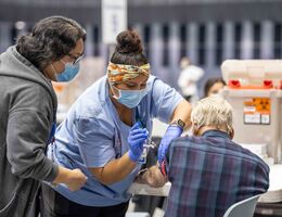 Nurse in blue scrubs giving a vaccine to a senior citizen as someone else looks on