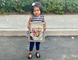 LLUCH patient, Lydia Cuasito, 3, holds a photo of herself taken in 2021 while undergoing treatment for a heart condition as an infant. Little girl wearing black and white dress with leggings and flats holds a photo of herself when she was in the NICU