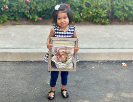 Little girl wearing black and white dress with leggings and flats holds a photo of herself when she was in the NICU