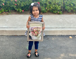 Little girl wearing black and white dress with leggings and flats holds a photo of herself when she was in the NICU