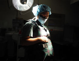Urology resident Ruby Kuang, MD, a contributing author and pregnant surgeon, was featured on the cover of the Journal of Urology. pregnant, female physician wearing a surgical mask and hair cap in a dark room under a light looking down at her belly
