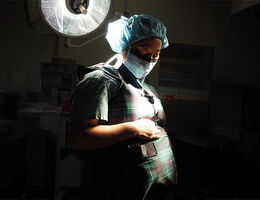 pregnant, female physician wearing a surgical mask and hair cap in a dark room under a light looking down at her belly