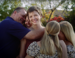 family surrounds teenage daughter, all in a group hug outside smiling broadly