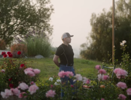 Young hispanic stands in garden with arms relaxed behind his back.
