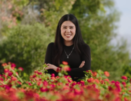 Young woman with dark hair and a black shirt stands smiling with her arms crossed in front. Red flowered bushes are in the foreground. 