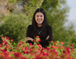 Young woman with dark hair and a black shirt stands smiling with her arms crossed in front. Red flowered bushes are in the foreground. 