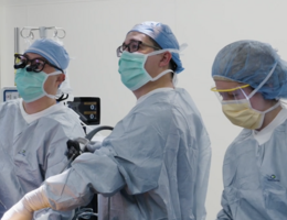 Three people in blue scrubs stand over operating table looking to the viewers left.