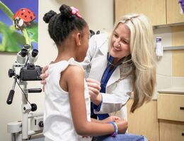 A healthcare provider wearing a white coat uses a stethoscope to examine a child seated on an exam table in a pediatric clinic. Medical equipment and cabinets are visible in the background.